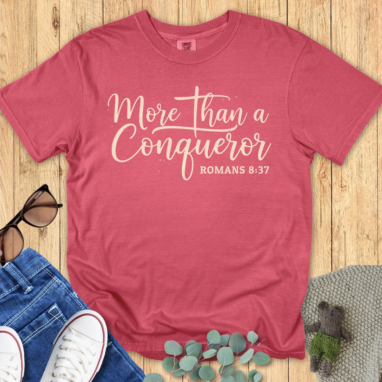 Flatlay of a Comfort Colors 1717 crimson “More Than a Conqueror – Romans 8:37” t-shirt featuring cream script typography. A vibrant women’s scripture tee — uplifting faith-based apparel for women and perfect Christian mom gift idea inspired by Romans 8:37.