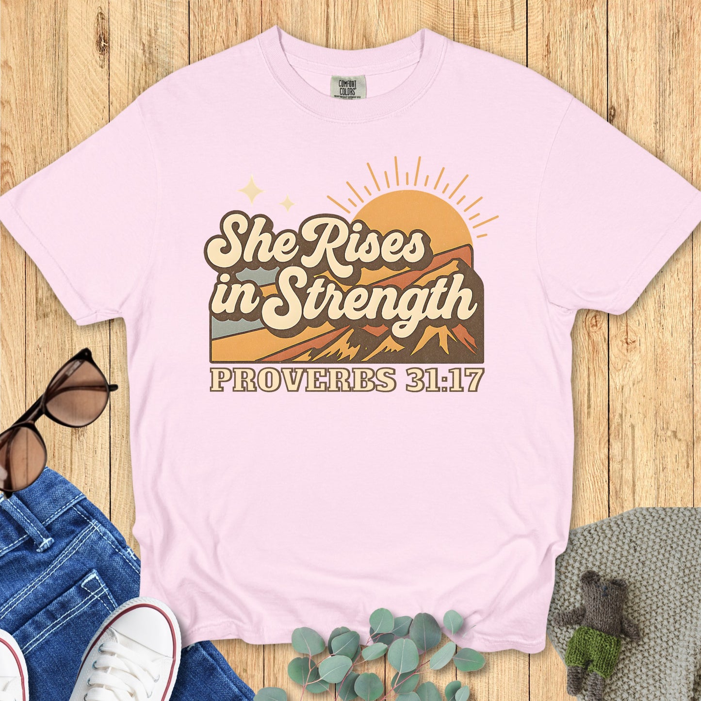 Comfort Colors 1717 blossom “She Rises in Strength – Proverbs 31:17” t-shirt in pastel pink with retro mountain sunrise artwork. A feminine women’s scripture tee — inspiring faith-based apparel for women and thoughtful Christian mom gift idea.