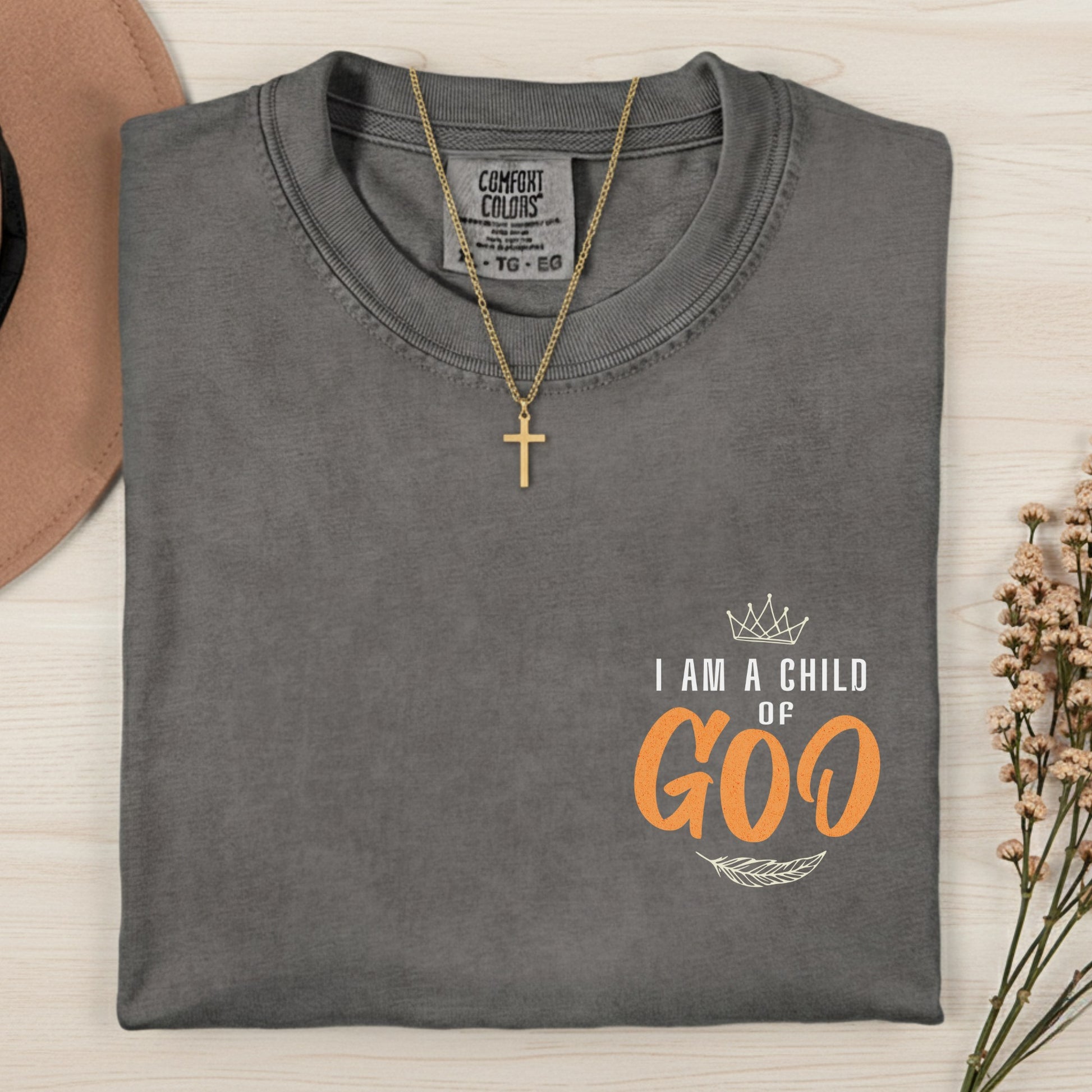 Flatlay of a Comfort Colors 1717 pepper “I Am a Child of God” t-shirt with orange lettering, crown, and feather design beside a cross necklace. A bold women’s scripture tee — inspiring faith-based apparel for women and modern Christian shirt, perfect Christian mom gift idea.