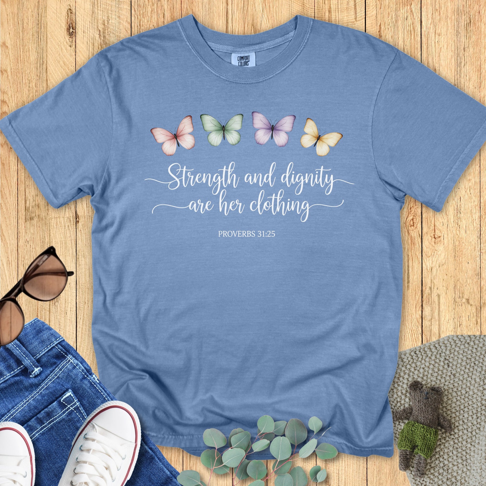 Flatlay of a Comfort Colors 1717 blue jean “Strength and dignity are her clothing – Proverbs 31:25” t-shirt featuring pastel butterflies and elegant script text. A beautiful women’s scripture tee — soft faith-based apparel for women and perfect Christian mom gift idea.