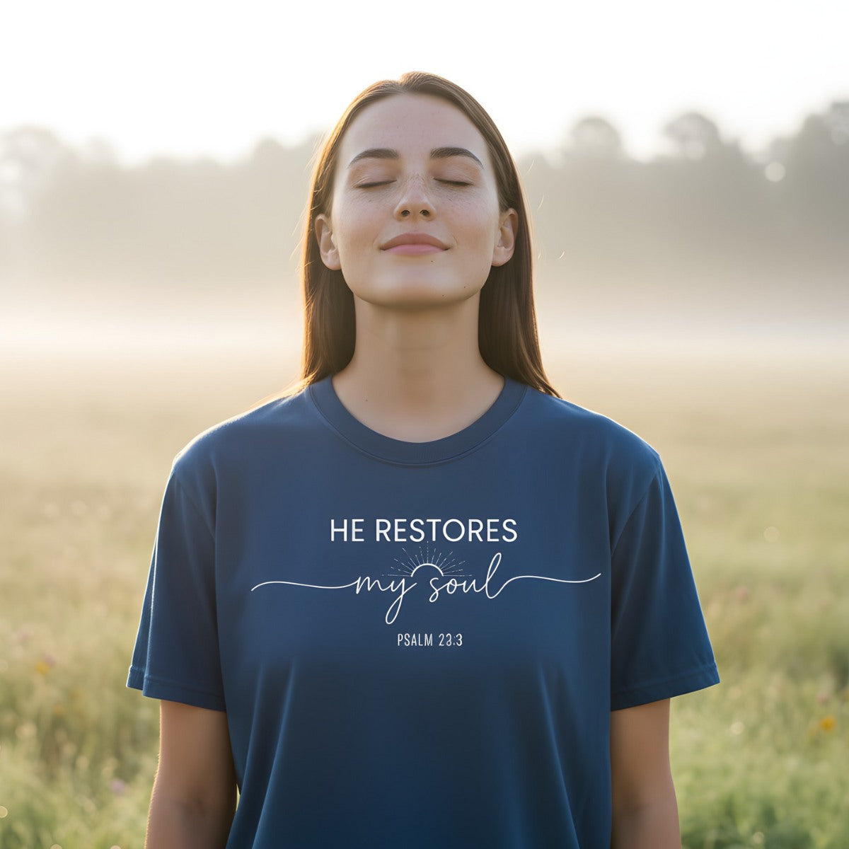 Woman standing peacefully in a misty morning field wearing a Comfort Colors 1717 midnight blue “He Restores My Soul – Psalm 23:3” t-shirt with sunrise line art. A serene women’s scripture tee — inspiring faith-based apparel for women and modern Christian shirt, perfect Christian mom gift idea.