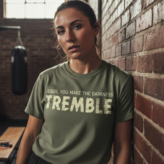 Woman standing against a brick wall in a rustic gym setting wearing a Comfort Colors 1717 sage “Jesus, You Make the Darkness Tremble” t-shirt. A bold women’s scripture tee — empowering faith-based apparel for women and modern Christian shirt, inspiring Christian mom gift idea.