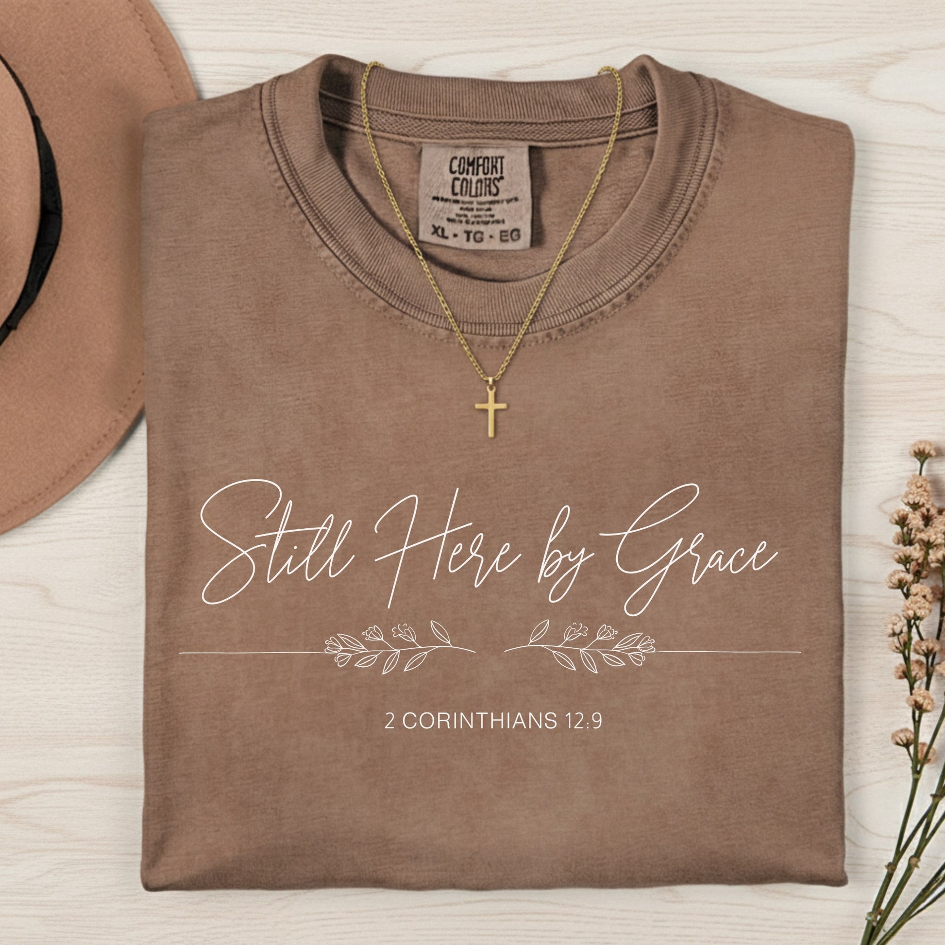 Flatlay of a Comfort Colors 1717 espresso “Still Here by Grace – 2 Corinthians 12:9” t-shirt featuring elegant cursive text and leaf accents. A soft women’s scripture tee — encouraging faith-based apparel for women and modern Christian shirt, meaningful Christian mom gift idea.
