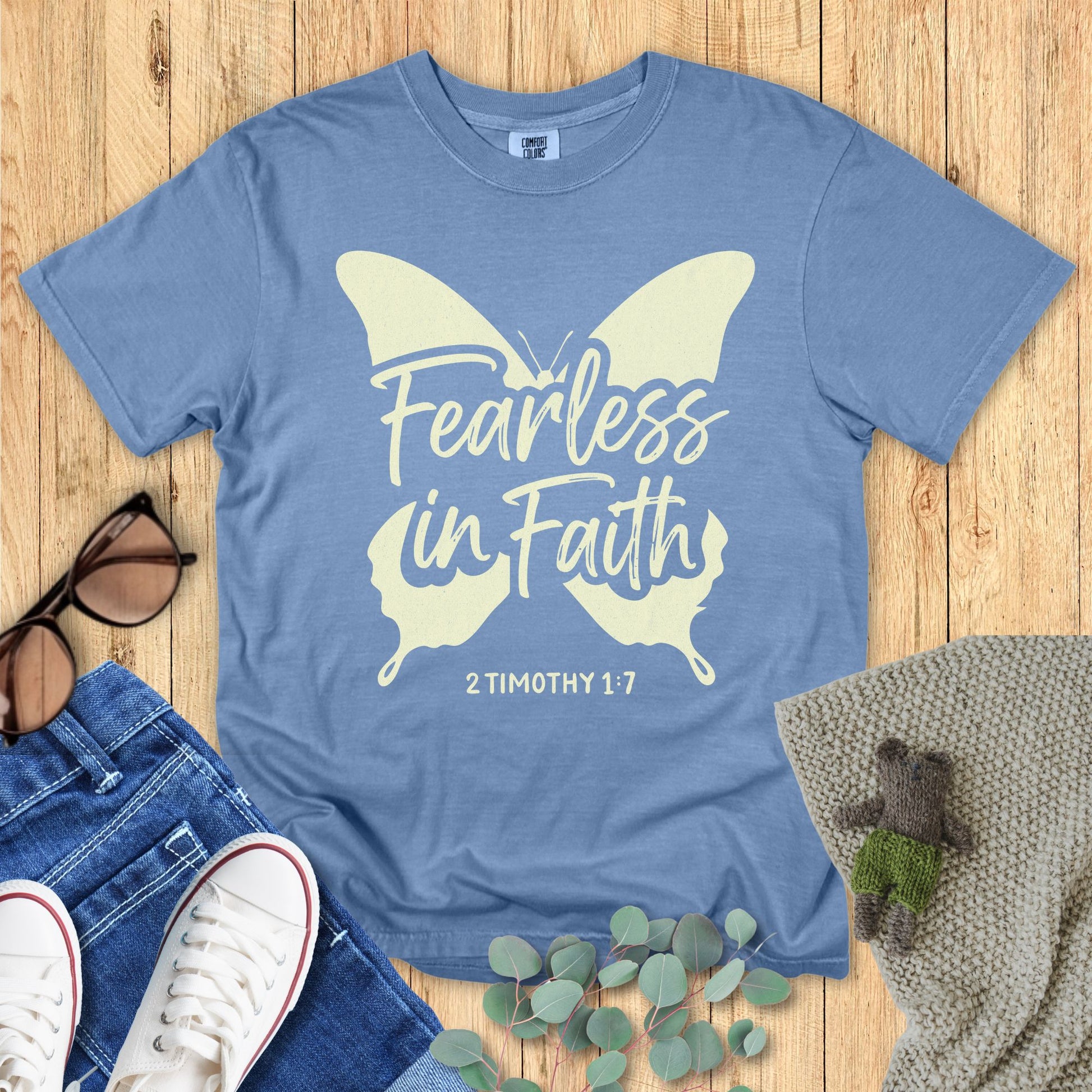 Flatlay of a Comfort Colors 1717 blue jean “Fearless in Faith – 2 Timothy 1:7” t-shirt with soft cream butterfly print and elegant lettering. A graceful women’s scripture tee — uplifting faith-based apparel for women and modern Christian shirt, lovely Christian mom gift idea.
