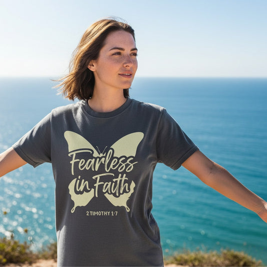 Woman standing by the ocean wearing a Comfort Colors 1717 pepper “Fearless in Faith – 2 Timothy 1:7” t-shirt with a butterfly design. A bold women’s scripture tee — empowering faith-based apparel for women and modern Christian shirt, meaningful Christian mom gift idea.