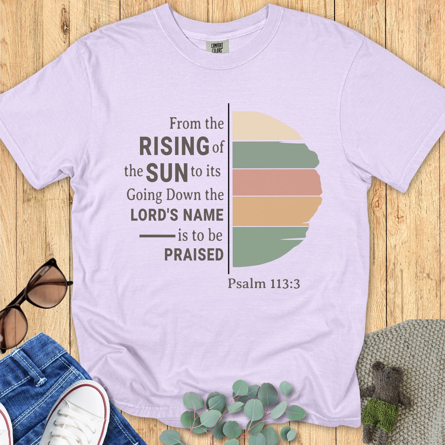 Flatlay of a Comfort Colors 1717 orchid “From the Rising of the Sun to its Going Down, the Lord’s Name is to be Praised – Psalm 113:3” t-shirt with muted sunrise graphic. A soft and inspiring women’s scripture tee — faith-based apparel for women and modern Christian shirt, lovely Christian mom gift idea.