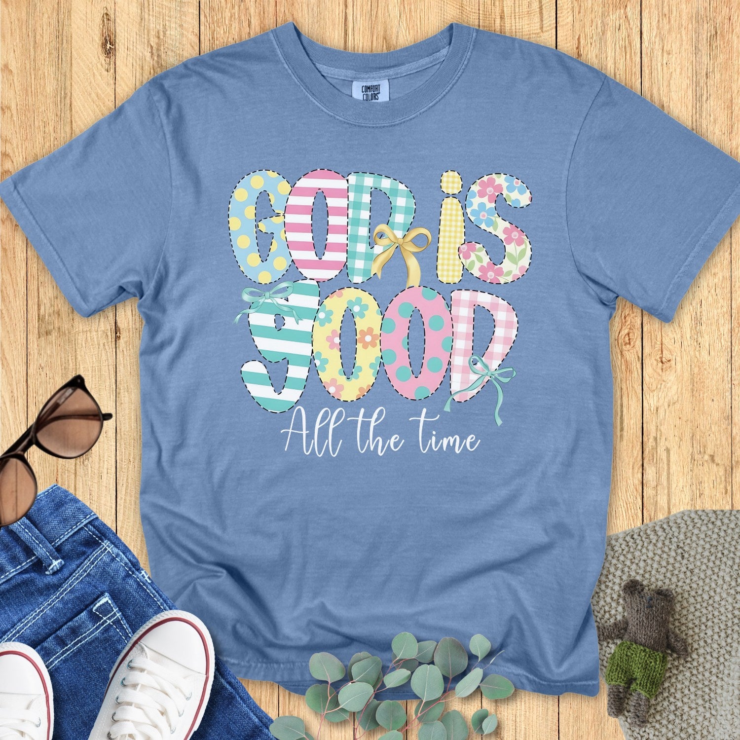 Flatlay of a Comfort Colors 1717 blue jean “God Is Good All the Time” t-shirt featuring pastel patchwork lettering with playful bow accents. A bright and joyful women’s scripture tee — faith-based apparel for women and modern Christian shirt, ideal Christian mom gift idea.