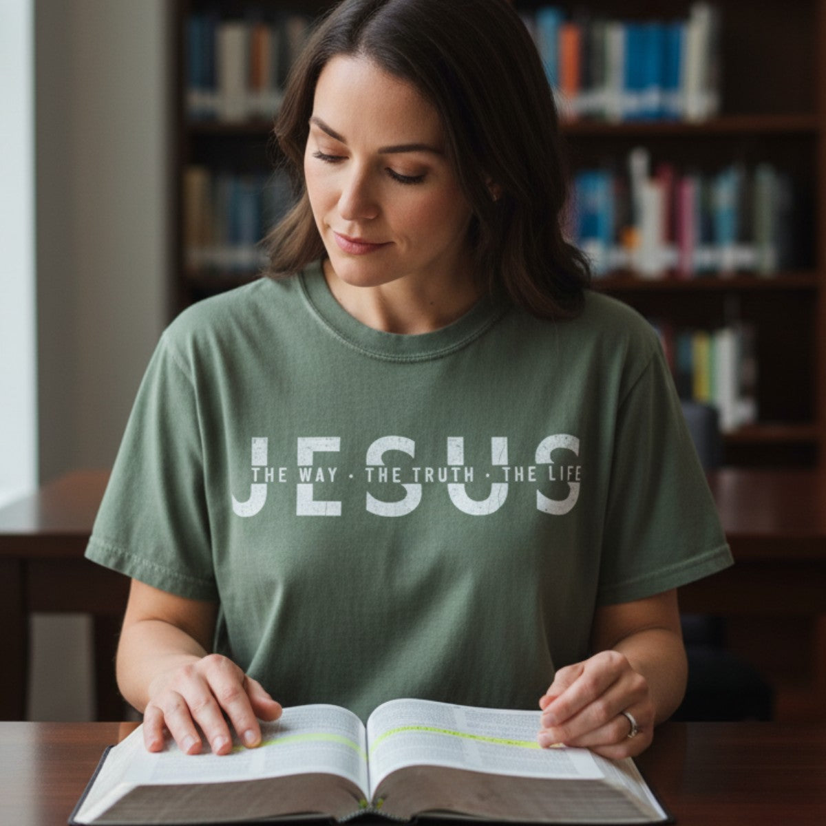 Woman reading her Bible in a library while wearing a Comfort Colors 1717 moss “Jesus — The Way, The Truth, The Life” t-shirt. A classic women’s scripture tee — faith-based apparel for women and modern Christian shirt from John 14:6, thoughtful Christian mom gift idea.