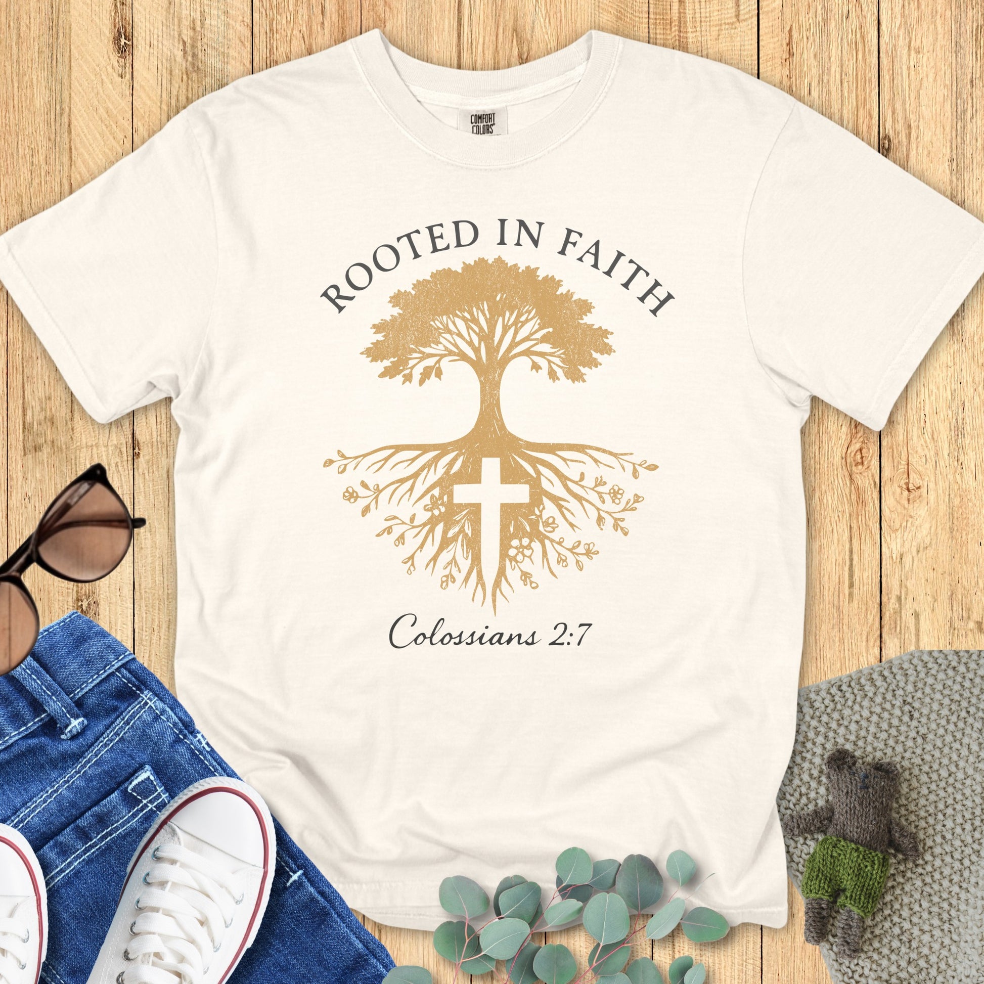 Flatlay of a Comfort Colors 1717 ivory “Rooted in Faith” t-shirt featuring a golden tree and cross design with Colossians 2:7 verse. A soft women’s scripture tee — faith-based apparel for women and modern Christian shirt, meaningful Christian mom gift idea.
