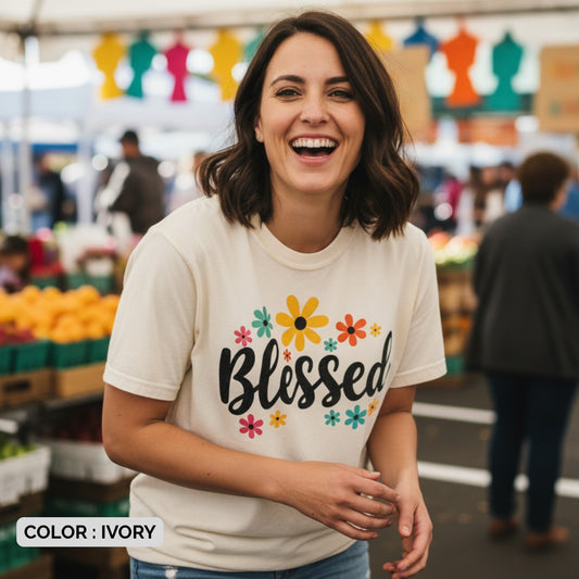 Woman smiling at an outdoor market wearing a Comfort Colors 1717 ivory βBlessedβ t-shirt with floral design. A casual womenβs scripture tee β faith-based apparel for women and modern Christian shirt, meaningful Christian mom gift idea.
