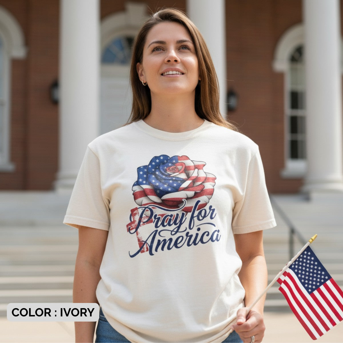 Woman standing proudly outdoors holding an American flag while wearing a Comfort Colors 1717 ivory “Pray for America” t-shirt. A meaningful women’s scripture tee — faith-based apparel for women and modern Christian shirt, uplifting Christian mom gift idea.