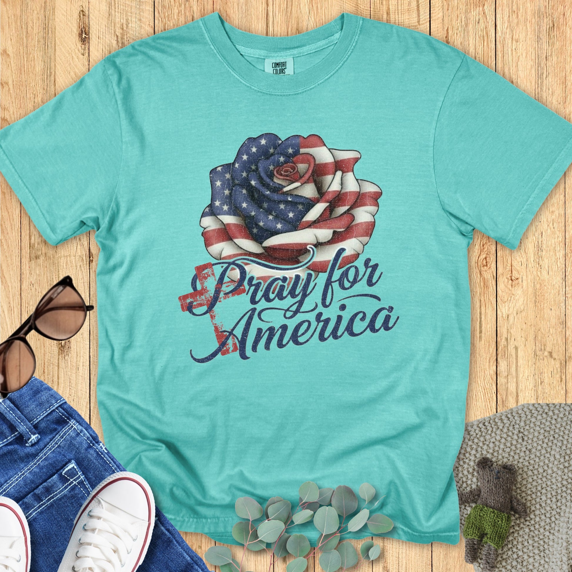 Flatlay of a Comfort Colors 1717 seafoam “Pray for America” t-shirt featuring a patriotic rose wrapped in the American flag. A bold women’s scripture tee — faith-based apparel for women and modern Christian shirt, perfect Christian mom gift idea for Independence Day or prayer gatherings.
