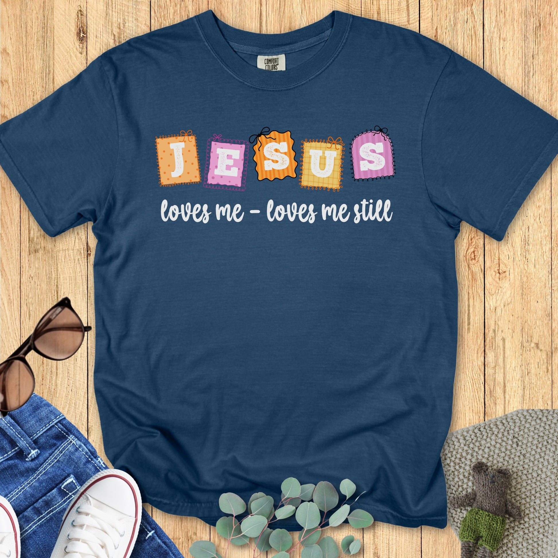 Flatlay of a Comfort Colors 1717 midnight blue “Jesus loves me – loves me still” t-shirt with vibrant orange, pink, and yellow stitched block letters. A playful women’s scripture tee — faith-based apparel for women and modern Christian shirt, uplifting Christian mom gift idea.
