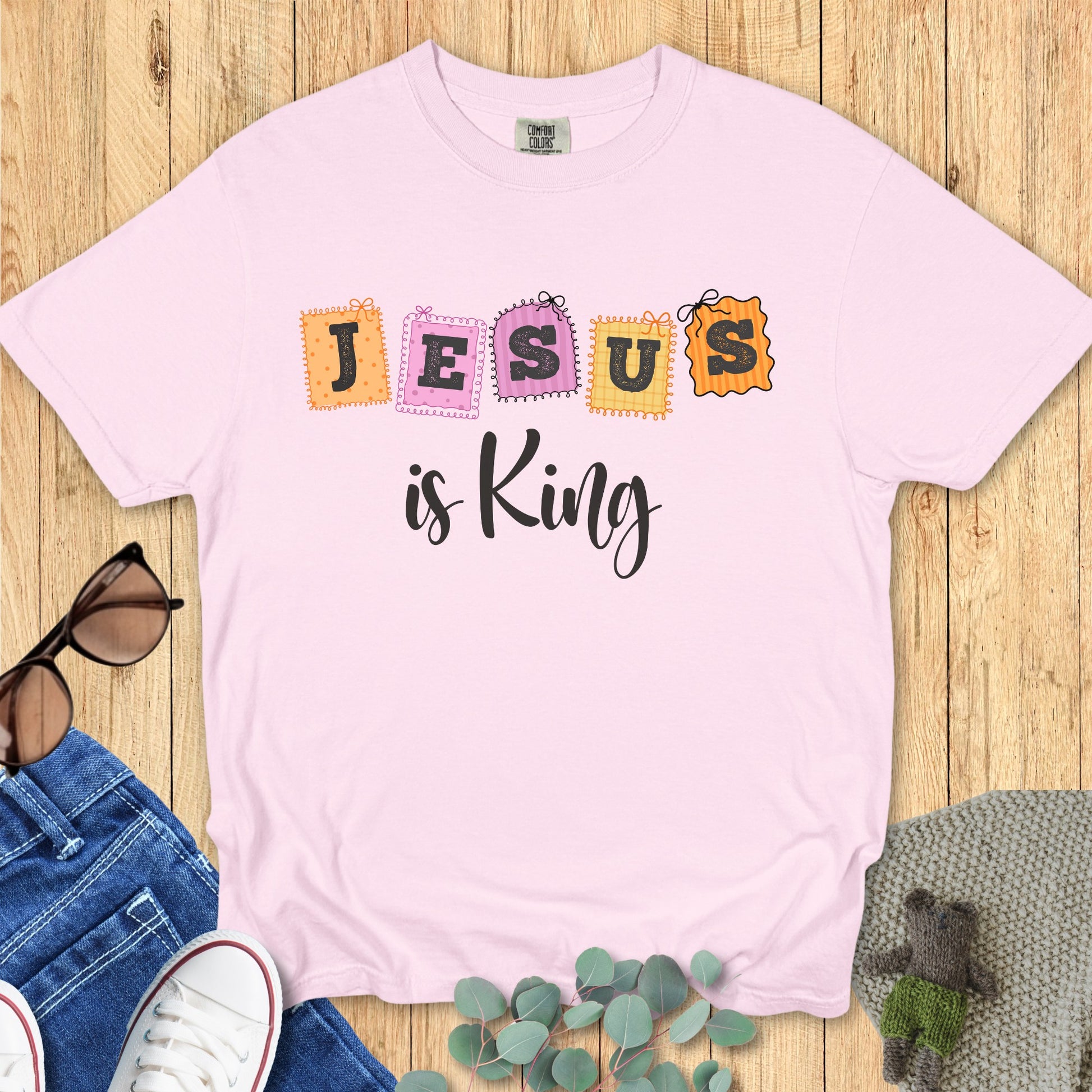 Flatlay of a Comfort Colors 1717 blossom pink “Jesus is King” t-shirt with vibrant patchwork-style letters. A joyful women’s scripture tee - faith-based apparel for women and modern Christian shirt, uplifting Christian mom gift idea for every season.