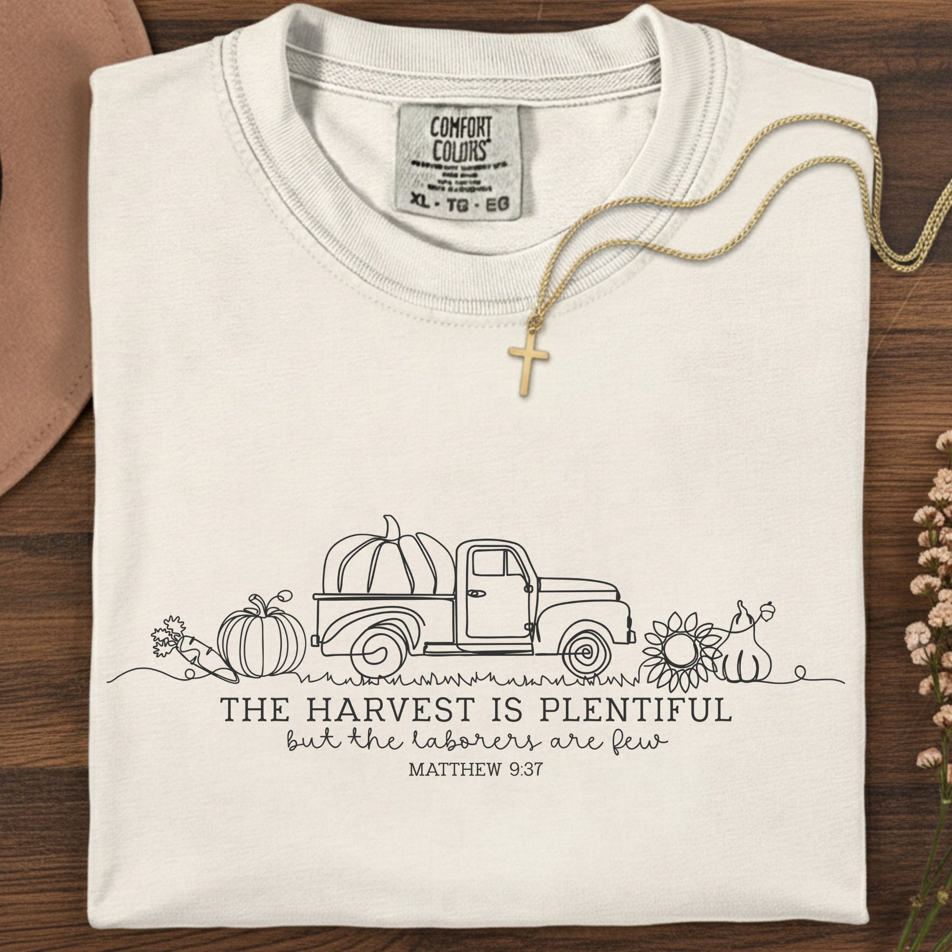 Folded Comfort Colors 1717 ivory “The harvest is plentiful but the laborers are few” t-shirt (Matthew 9:37) styled with gold cross necklace. A neutral women’s scripture tee -  faith-based apparel for women and modern Christian shirt, timeless Christian mom gift idea for the season.
