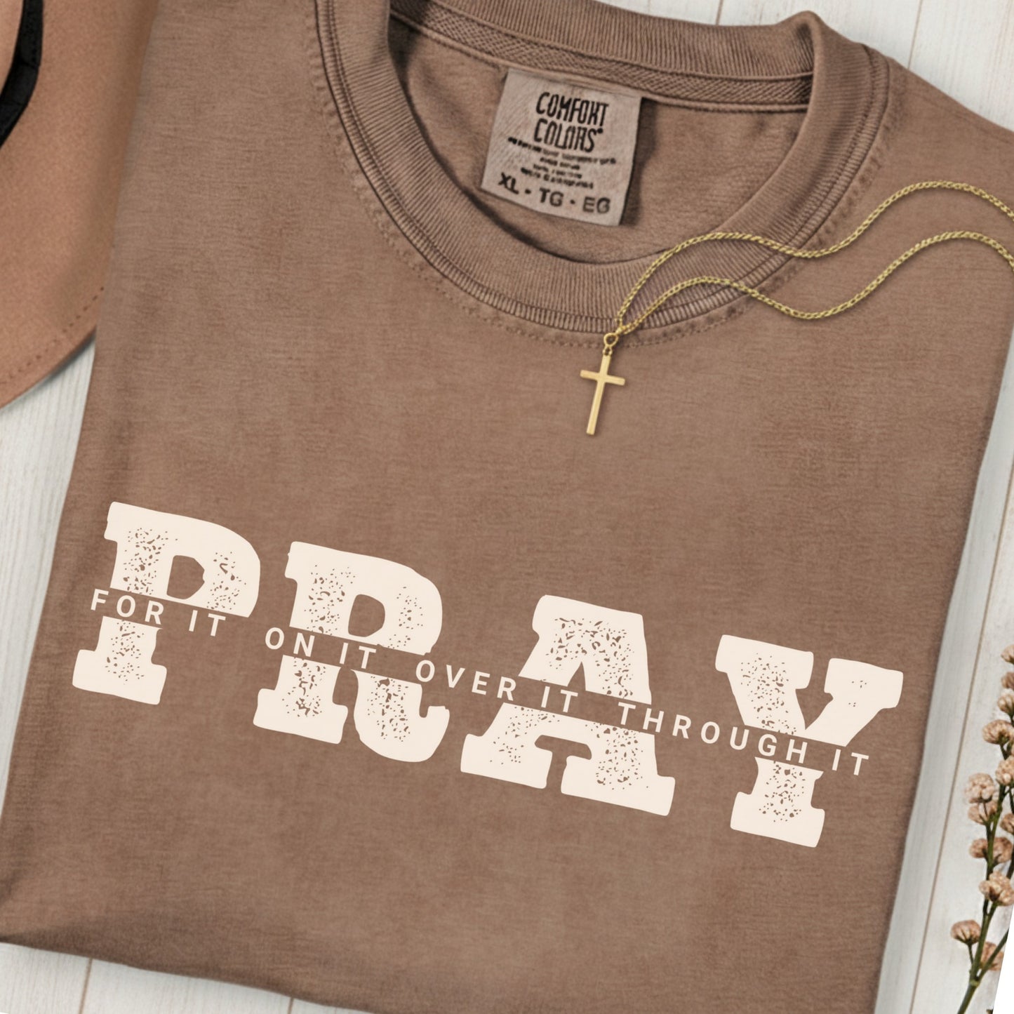 Folded Comfort Colors 1717 espresso “PRAY for it, on it, over it, through it” t-shirt with gold cross necklace on top. A classic women’s scripture tee -  faith-based apparel for women and modern Christian shirt, ideal Christian mom gift idea and prayer reminder.