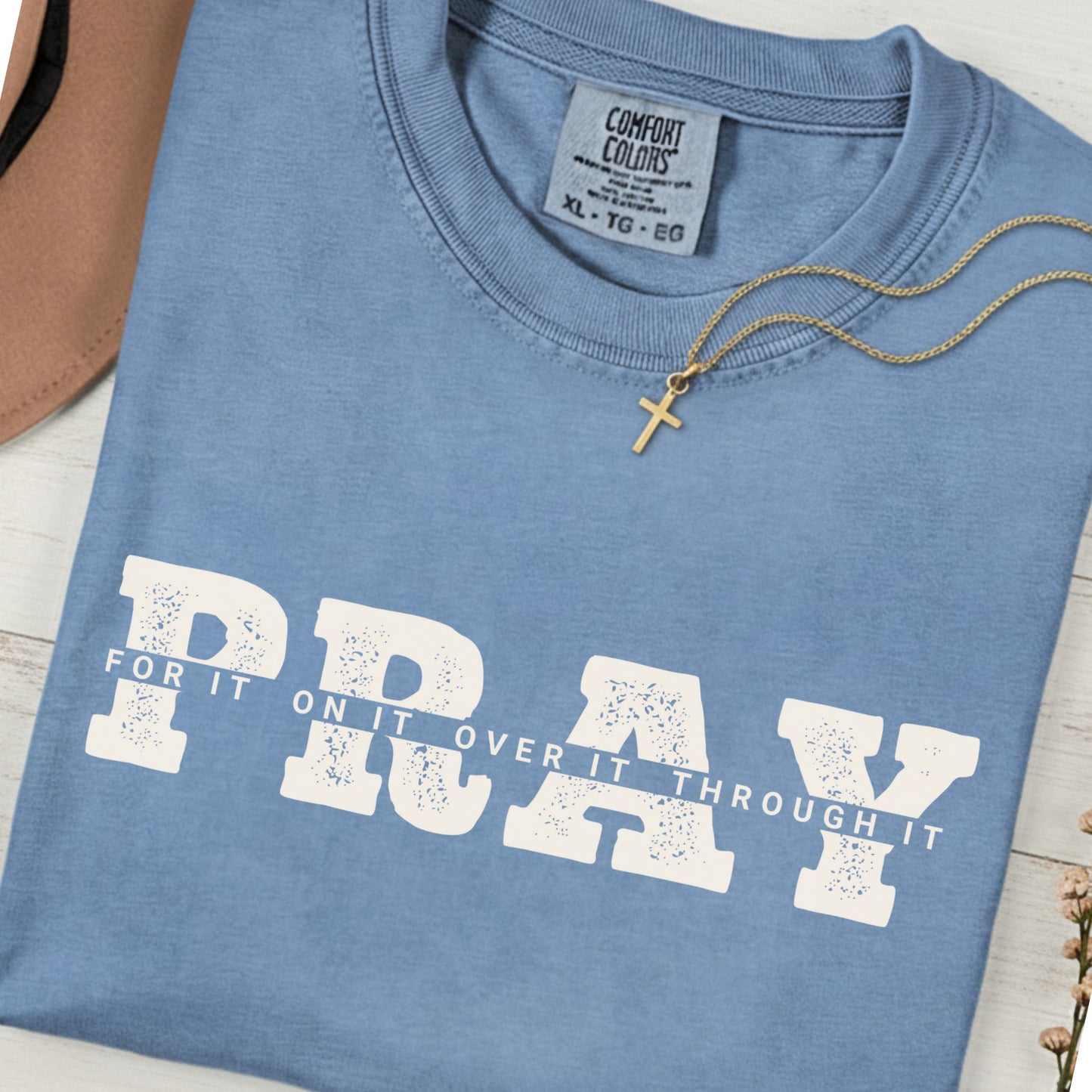 Folded Comfort Colors 1717 blue jean “PRAY for it, on it, over it, through it” t-shirt styled with a gold cross necklace. A soft women’s scripture tee -  faith-based apparel for women and modern Christian shirt, uplifting Christian mom gift idea for everyday faith wear.