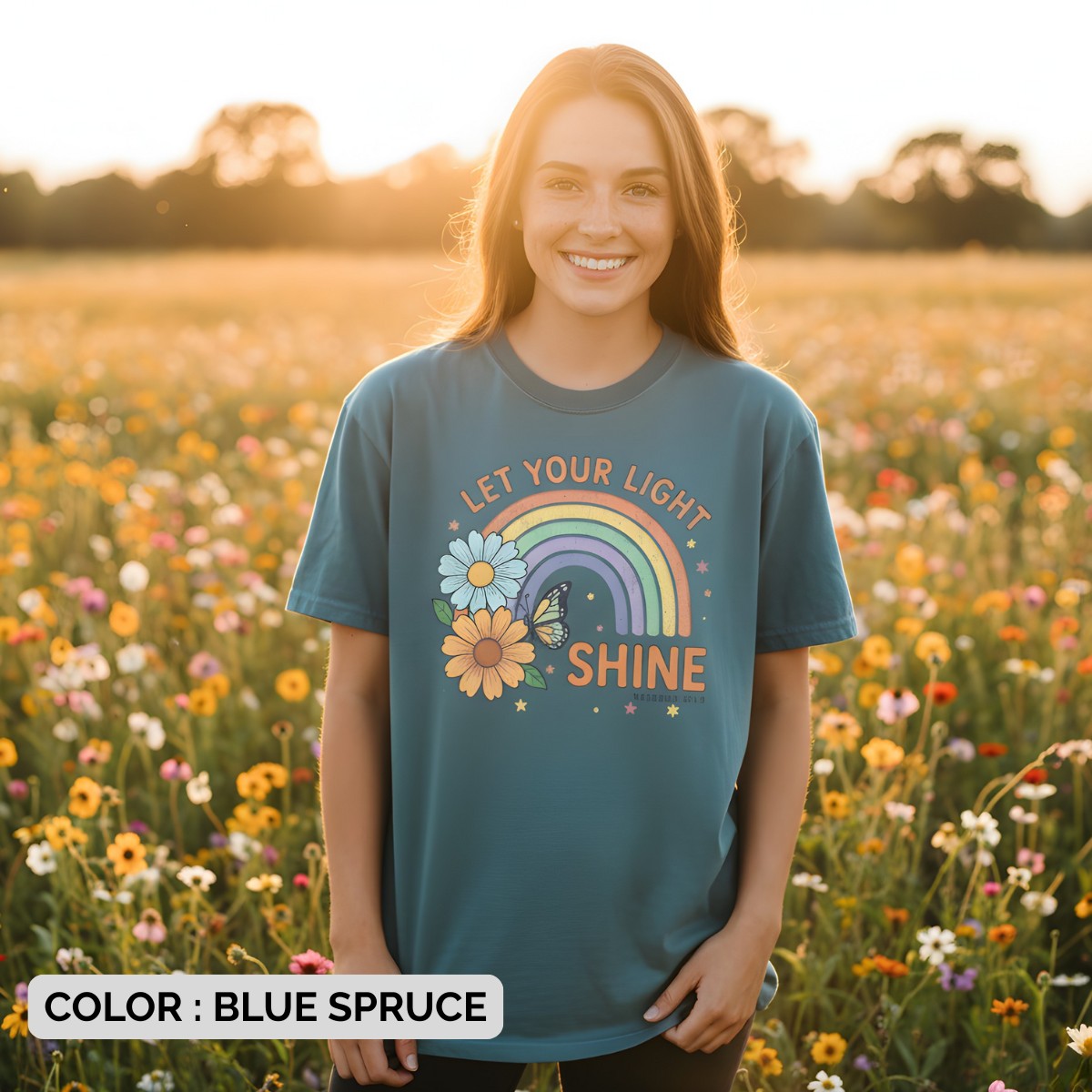 Woman smiling in a flower field wearing a Comfort Colors 1717 blue spruce “Let Your Light Shine” Matthew 5:16 t-shirt featuring rainbow and daisy artwork. A cheerful women’s scripture tee - faith-based apparel for women and modern Christian shirt, uplifting Christian mom gift idea for everyday inspiration.