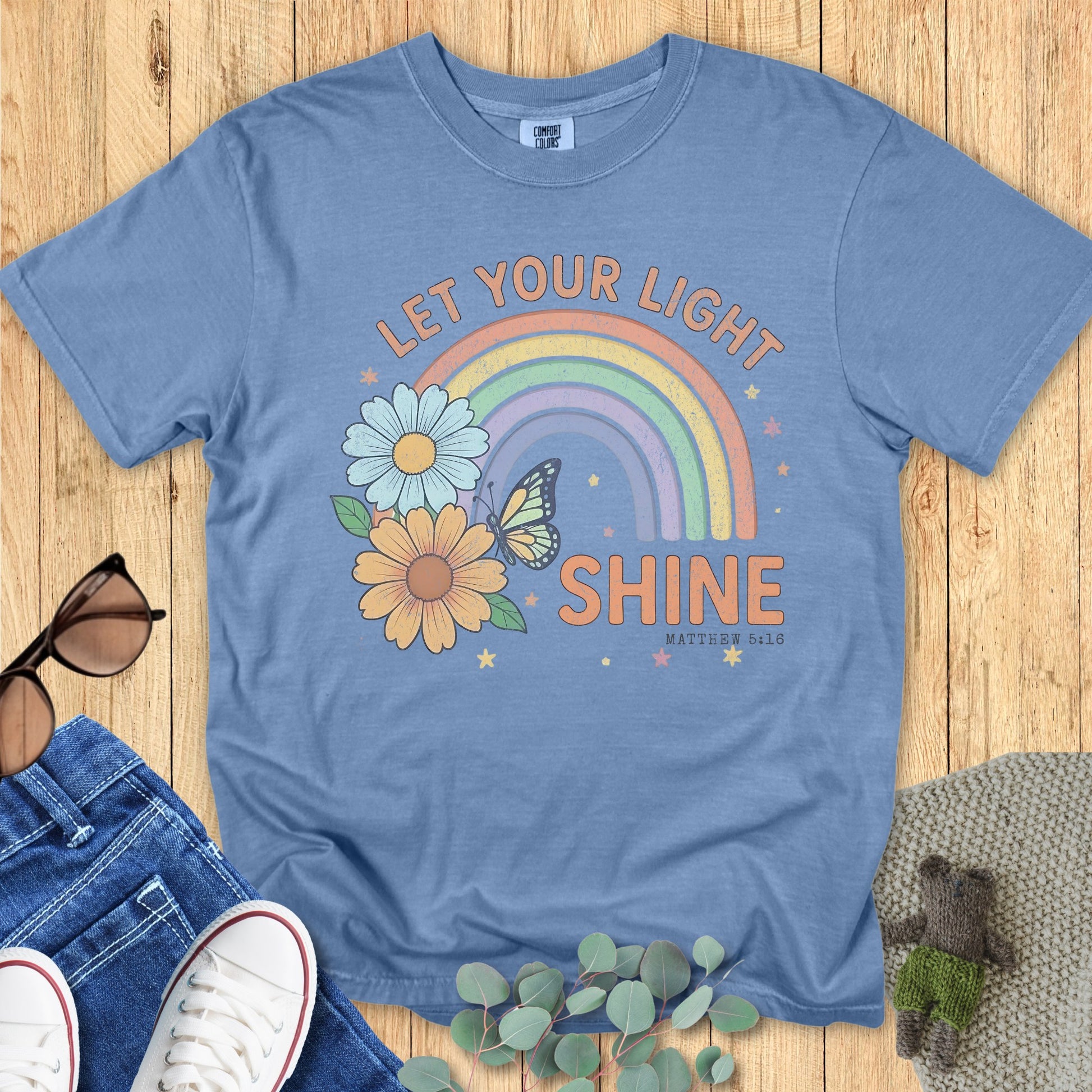 Flatlay of a Comfort Colors 1717 blue jean “Let Your Light Shine” Matthew 5:16 t-shirt with vintage pastel rainbow and flower graphic. A stylish women’s scripture tee - faith-based apparel for women and modern Christian shirt, thoughtful Christian mom gift idea that radiates positivity.