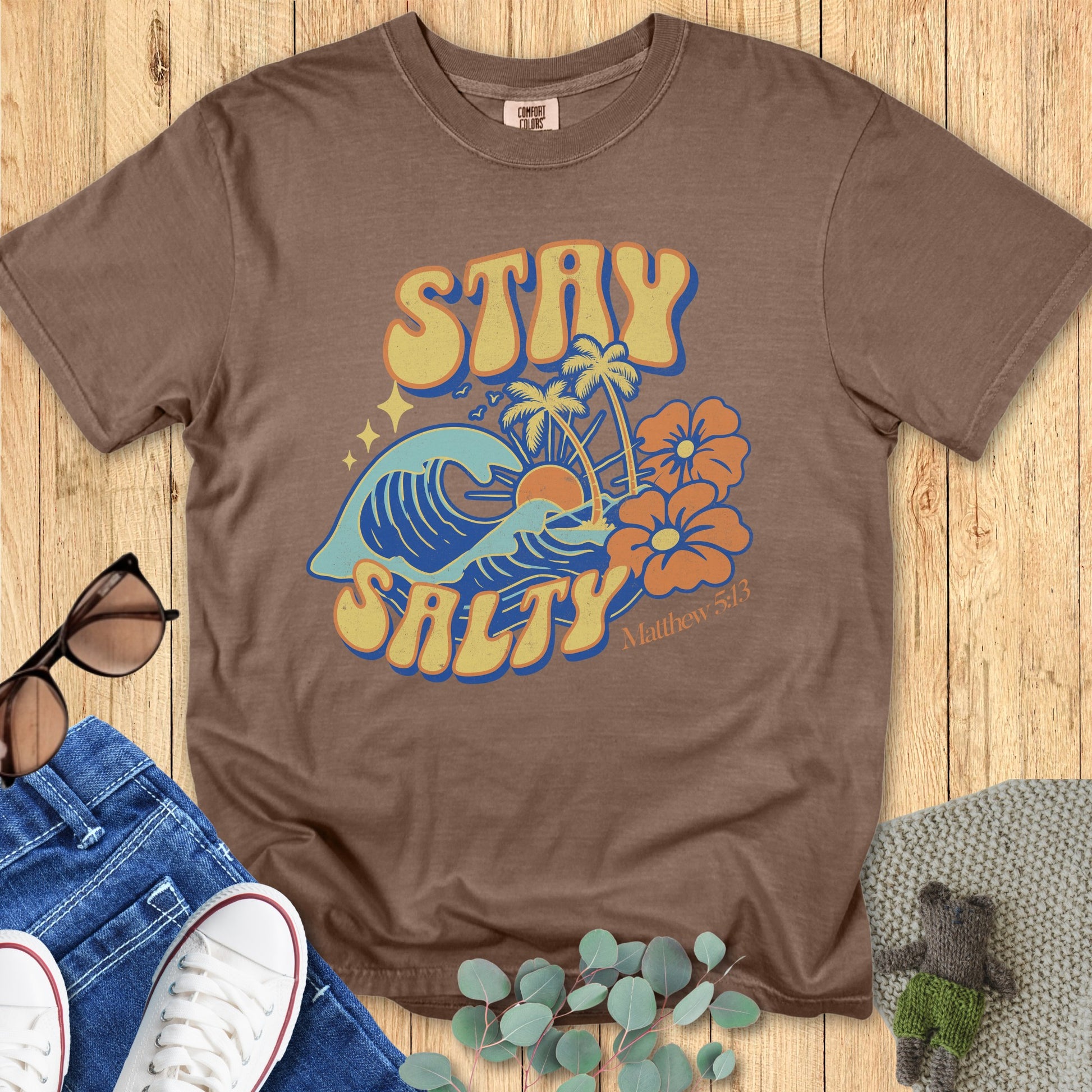 Flatlay of a Comfort Colors 1717 espresso “Stay Salty” t-shirt with tropical wave and sunset illustration and Matthew 5:13 verse. A stylish women’s scripture tee - faith-based apparel for women and modern Christian shirt, thoughtful Christian mom gift idea for beach lovers.