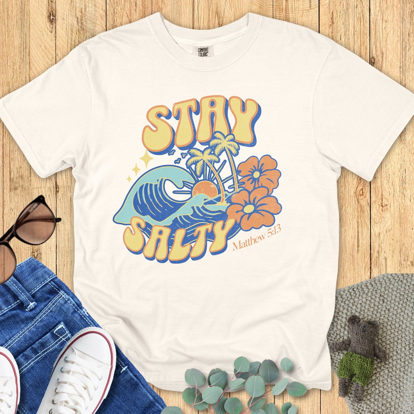 Flatlay of a Comfort Colors 1717 ivory “Stay Salty” t-shirt featuring vintage surf-style art with waves, sun, and hibiscus flowers inspired by Matthew 5:13. A cheerful women’s scripture tee - faith-based apparel for women and modern Christian shirt, perfect Christian mom gift idea for everyday faith and joy.