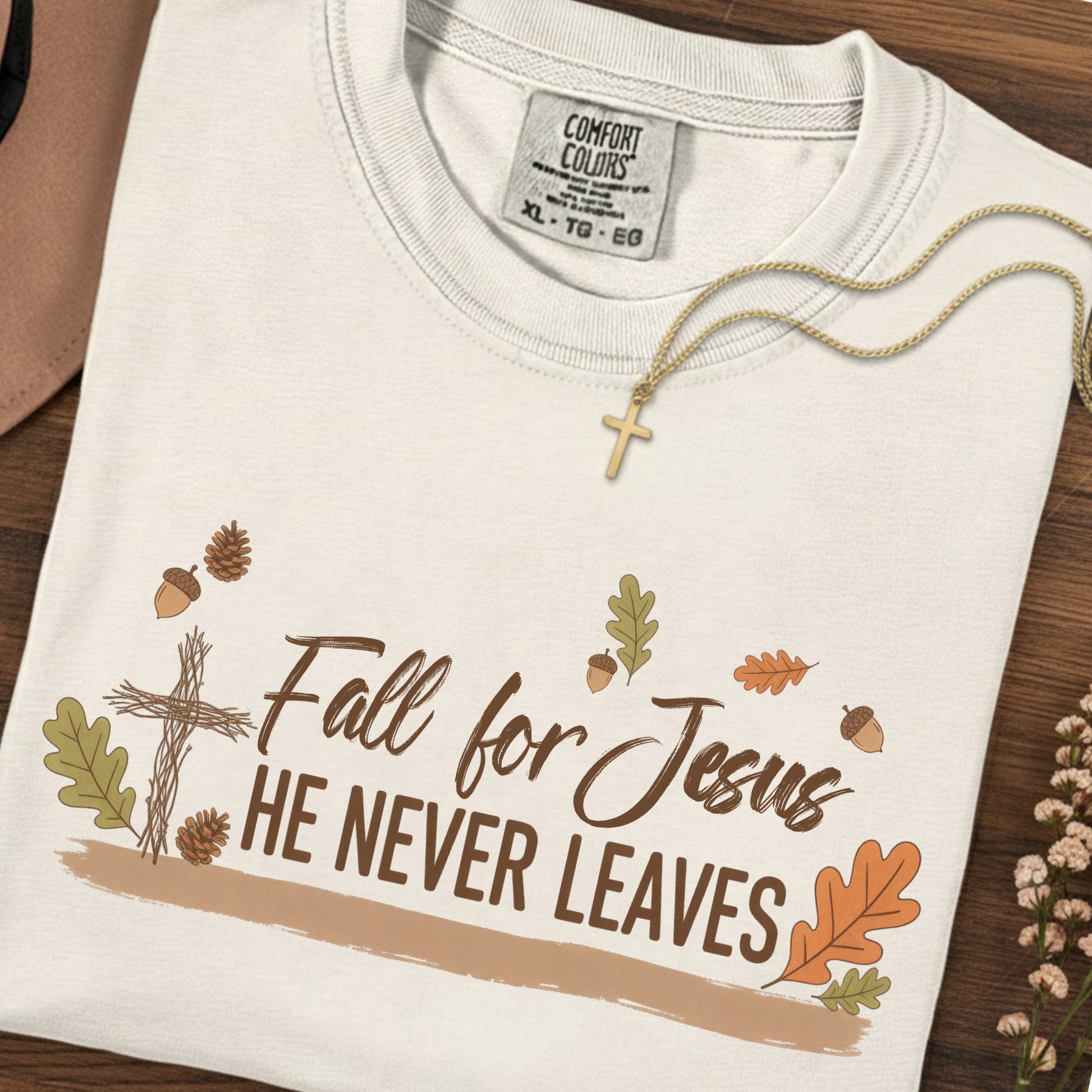 Flatlay of a Comfort Colors 1717 ivory “Fall for Jesus, He Never Leaves” t-shirt decorated with fall leaves, pinecones, and a twig cross. A warm women’s scripture tee -  faith-based apparel for women and modern Christian shirt, ideal Christian mom gift idea that blends faith and seasonal charm.