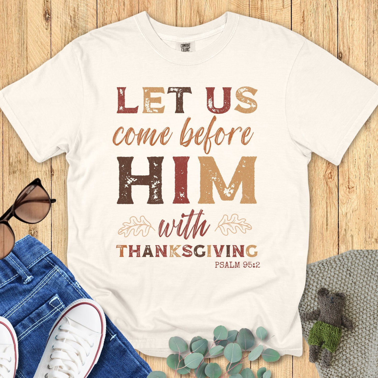Ivory Christian Thanksgiving shirt flat lay with the verse “Let Us Come Before Him With Thanksgiving, Psalm 95:2.” Distressed vintage fall lettering, oak leaf accents, faith-based fall outfit, Comfort Colors inspired.