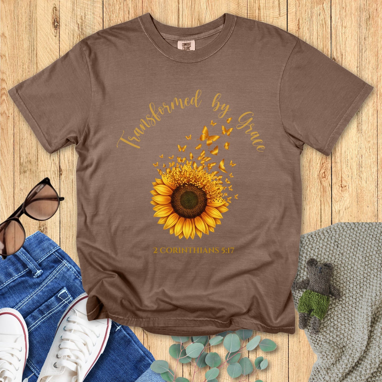 Brown t-shirt with sunflower design and text on a wooden surface with accessories.