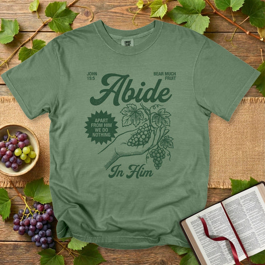 Comfort Colors 1717 hemp green Christian women tee featuring the “Abide in Him” John 15:5 design with a hand holding grape vines, styled in vintage faith wear. Inspirational Christian tshirt for US faith wear outfits, displayed flat-lay with grapes and an open Bible.