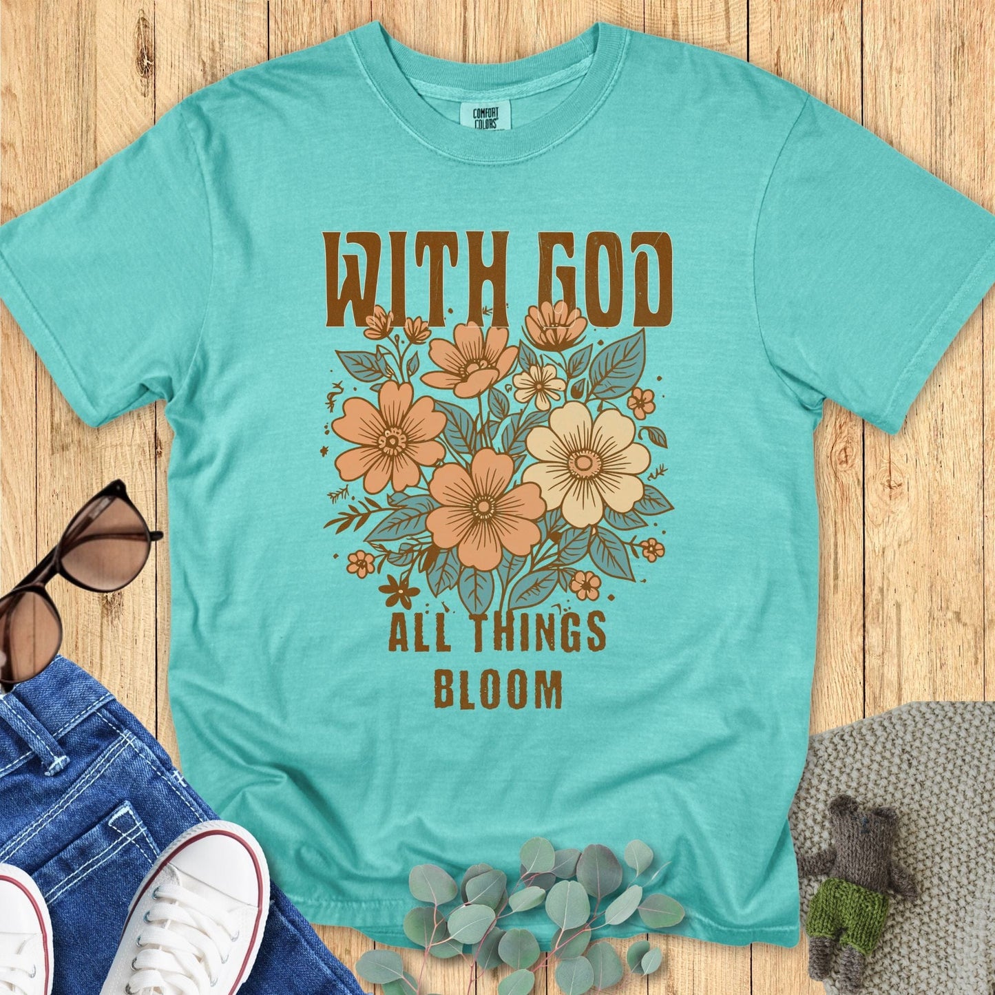 Christian women’s Comfort Colors 1717 t-shirt in Seafoam featuring peach floral artwork and text that reads ‘With God All Things Bloom.’ Shown in a styled flat lay with casual accessories
