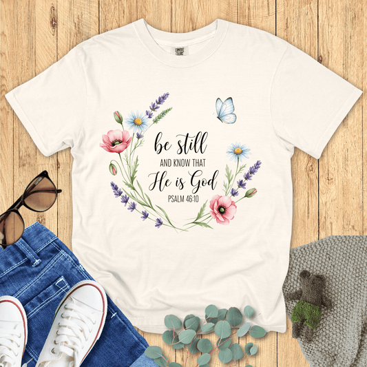 Ivory Comfort Colors Christian T-shirt with floral wreath and butterfly design featuring scripture "Be Still and Know That He Is God, Psalm 46:10." Inspirational Christian tee flat lay with jeans, sneakers, and sunglasses on wood background.