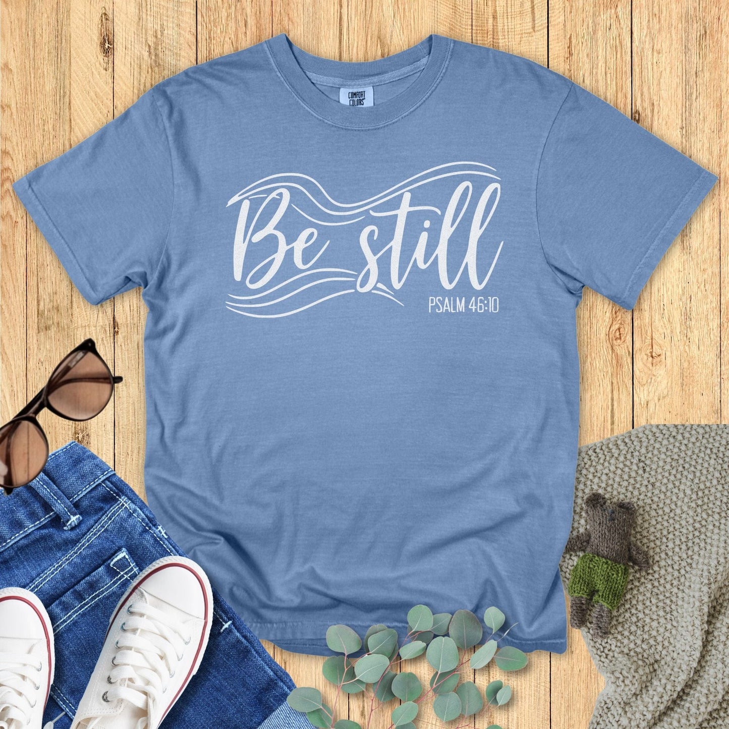Blue jean Christian T-shirt flat lay featuring the phrase “Be Still” with flowing white wave accents and “Psalm 46:10.” Displayed with sunglasses, denim jeans, sneakers, eucalyptus leaves, and a knitted blanket on a wooden background
