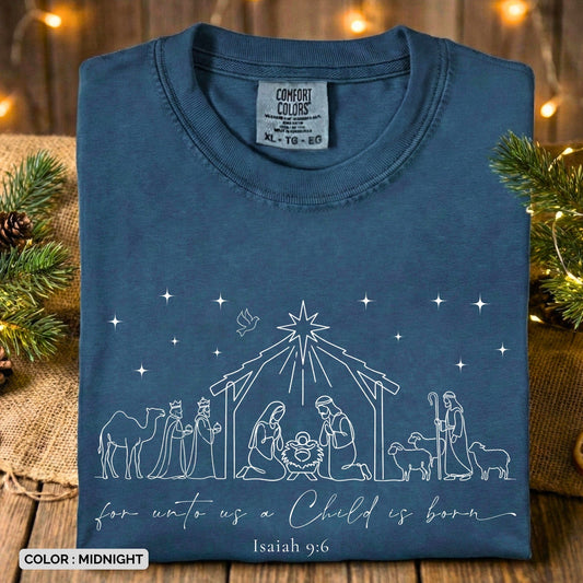 Folded Comfort Colors 1717 midnight Christian women tee with a delicate nativity line drawing, including the Holy Family, shepherds, wise men, and shining star, paired with “For unto us a Child is born – Isaiah 9:6.” Cozy Christmas US faith wear arranged with pine greenery and twinkling lights.