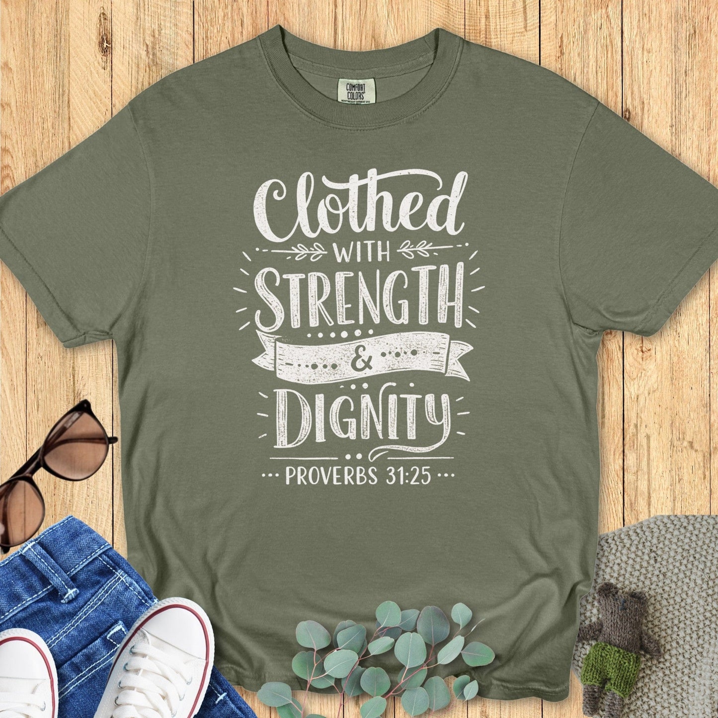 Flatlay of a Comfort Colors 1717 sage “Clothed with Strength & Dignity – Proverbs 31:25” t-shirt featuring elegant white typography with decorative flourishes. A soft, faith-based apparel piece for women — ideal Christian mom gift and daily encouragement shirt.