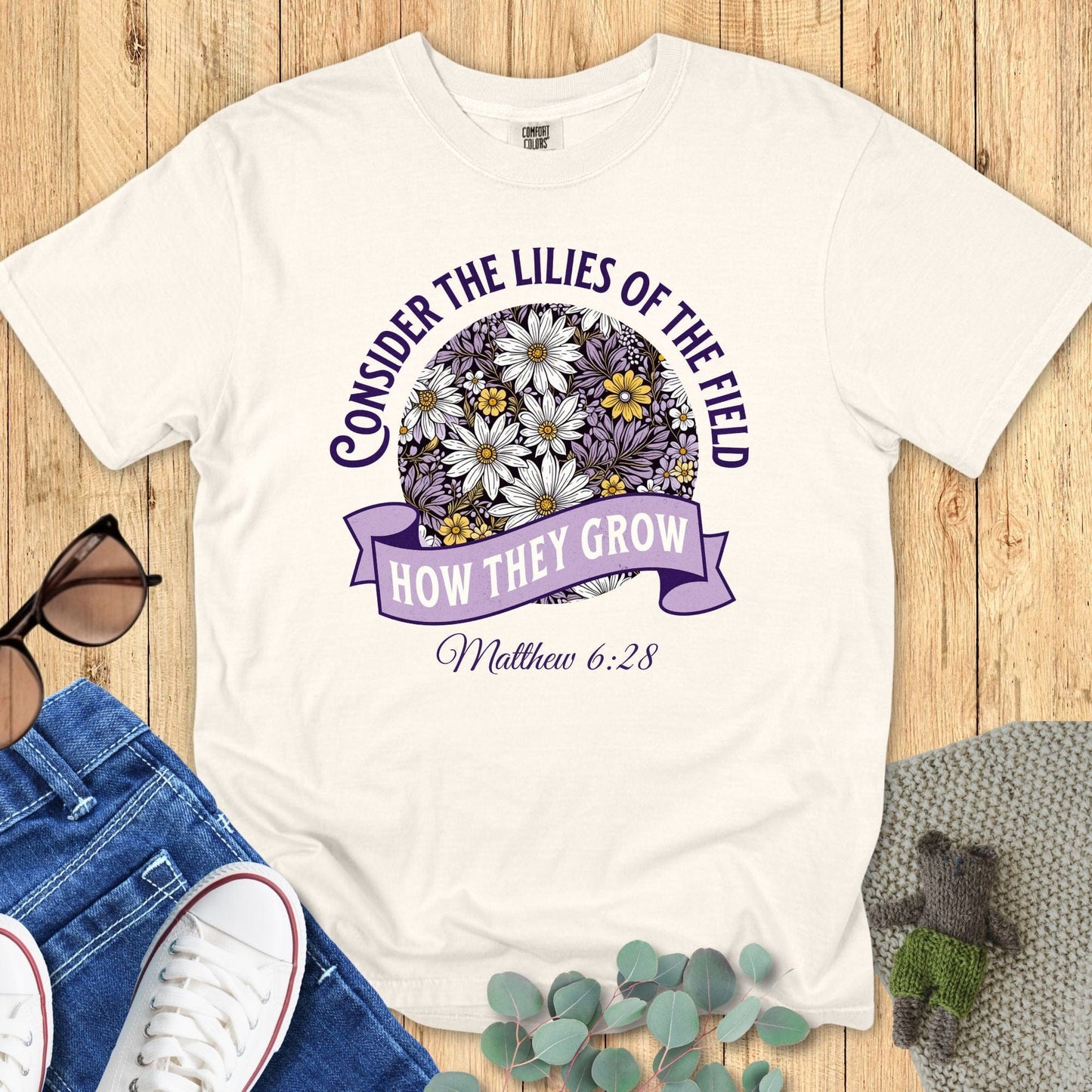 Comfort Colors 1717 Ivory Christian T-shirt featuring floral circle graphic with daisies and banner text “Consider the lilies of the field, how they grow – Matthew 6:28.” Flat lay with casual accessories.