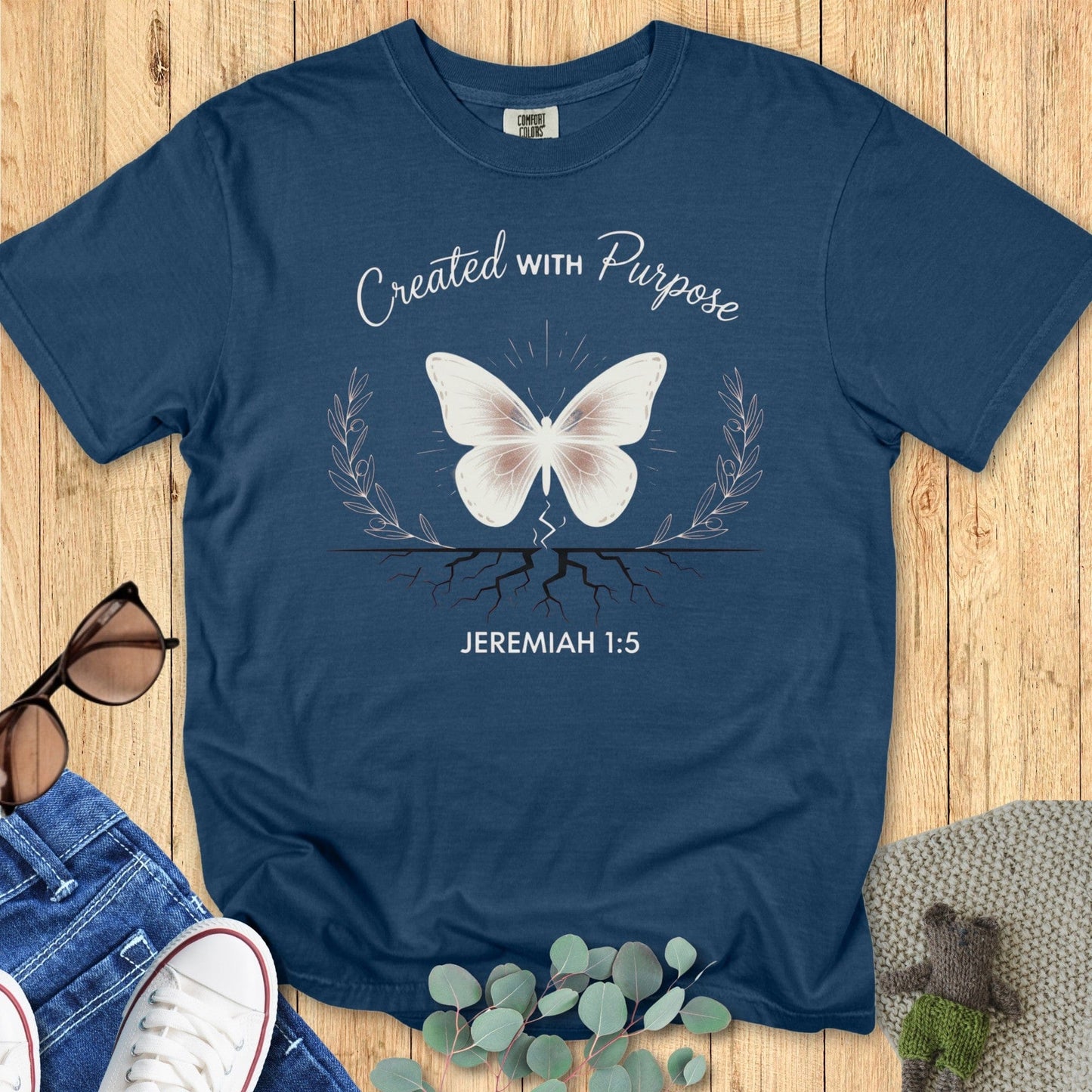 Flatlay of a Comfort Colors 1717 midnight “Created With Purpose – Jeremiah 1:5” t-shirt featuring a soft white butterfly emerging from roots with delicate branches. A meaningful women’s scripture tee — elegant faith-based apparel for women and a thoughtful Christian mom gift idea.