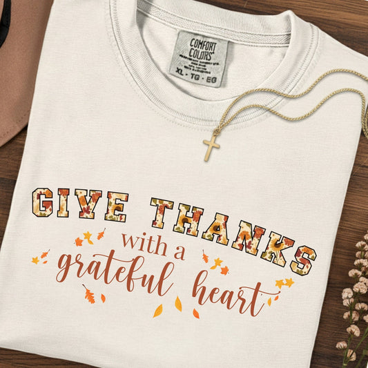 Folded Comfort Colors 1717 ivory “Give Thanks with a Grateful Heart” t-shirt with autumn-themed design and cross necklace beside it. A simple women’s scripture tee - faith-based apparel for women and modern Christian shirt, charming Christian mom gift idea for giving thanks in all seasons.