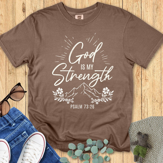 Comfort Colors 1717 espresso brown T-shirt featuring white design “God is my Strength – Psalm 73:26” with mountain, floral, and sunburst elements. Flat lay with casual lifestyle props on wood surface
