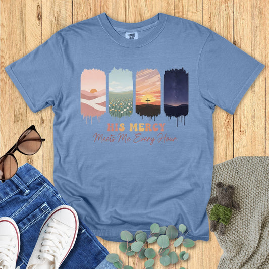 Comfort Colors Christian t-shirt in blue jean with Christian design “His Mercy Meets Me Every Hour.” Artwork shows four scenic panels—soft desert sunrise, floral field, cross at sunset, and starry night—symbolizing God’s mercy throughout the day. Flat lay mockup with denim, sneakers, and eucalyptus leave