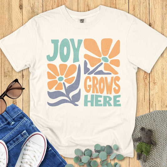 Flat lay of a Ivory T-shirt with the same “Joy Grows Here” floral design, styled on a wooden background alongside sunglasses, jeans, white sneakers, and greenery