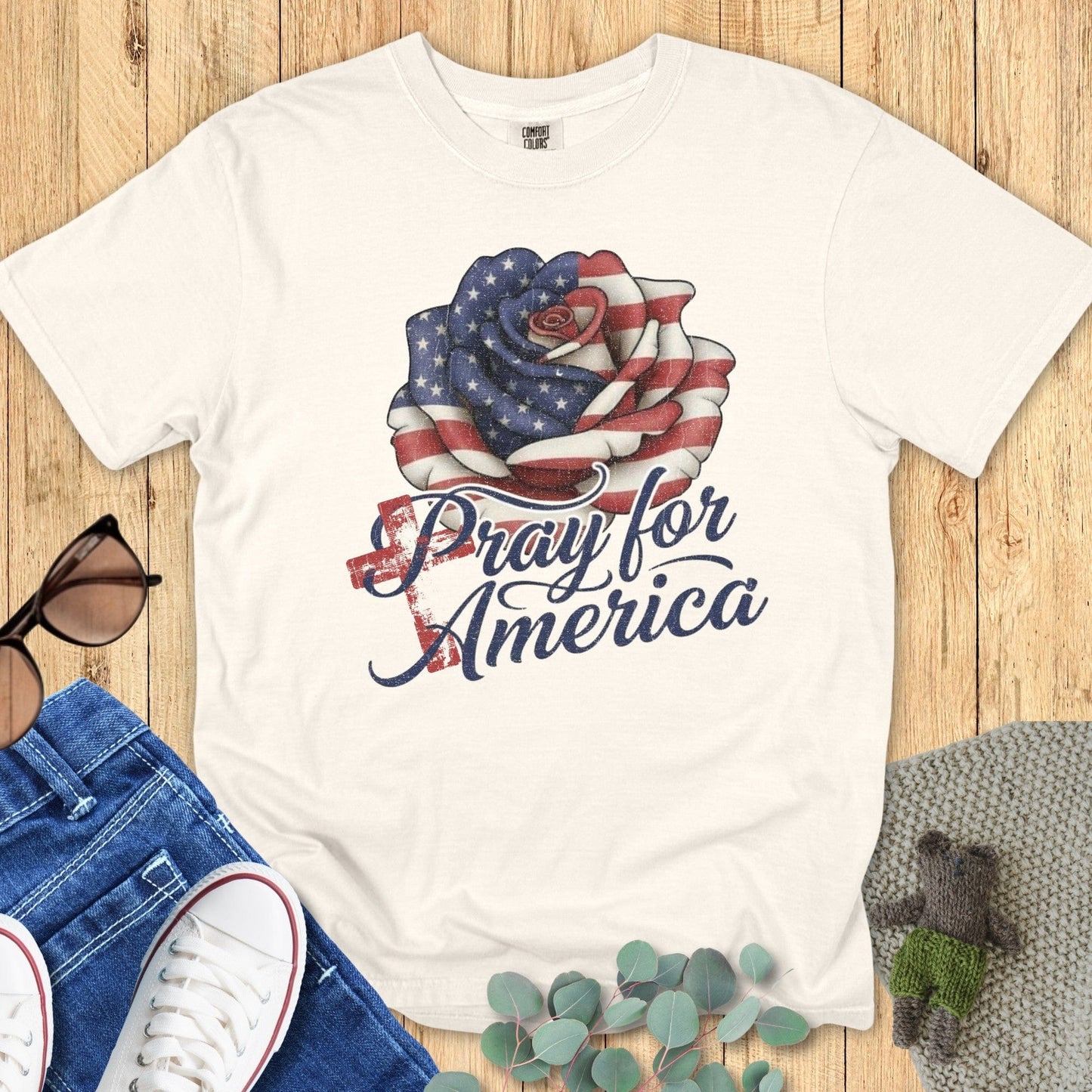 Flatlay of a Comfort Colors 1717 ivory “Pray for America” t-shirt featuring a distressed American flag rose design. A classic women’s scripture tee — faith-based apparel for women and modern Christian shirt, ideal Christian mom gift idea for prayer warriors and believers.