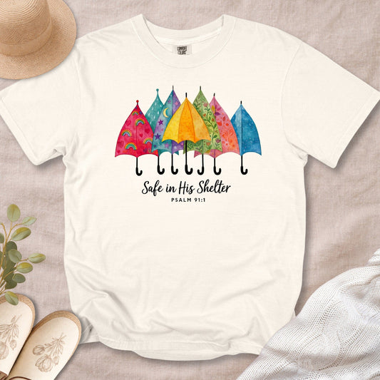 Flat lay of an Ivory Comfort Colors 1717 tshirt featuring colorful watercolor umbrellas. A cozy Christian women tee with "Safe in His Shelter" Psalm 91:1 scripture, perfect for US faith wear collections.