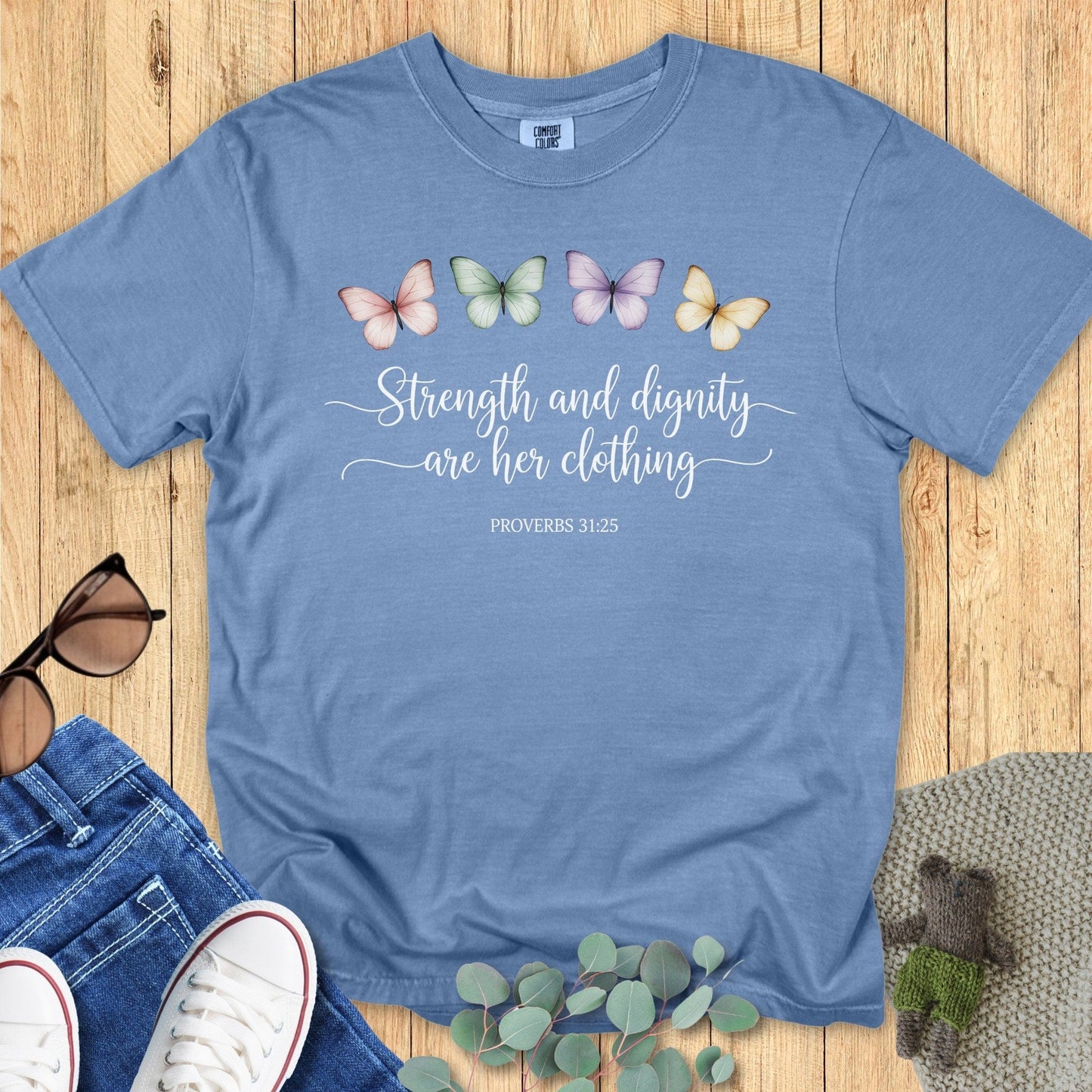 Flatlay of a Comfort Colors 1717 blue jean “Strength and dignity are her clothing – Proverbs 31:25” t-shirt featuring pastel butterflies and elegant script text. A beautiful women’s scripture tee — soft faith-based apparel for women and perfect Christian mom gift idea.