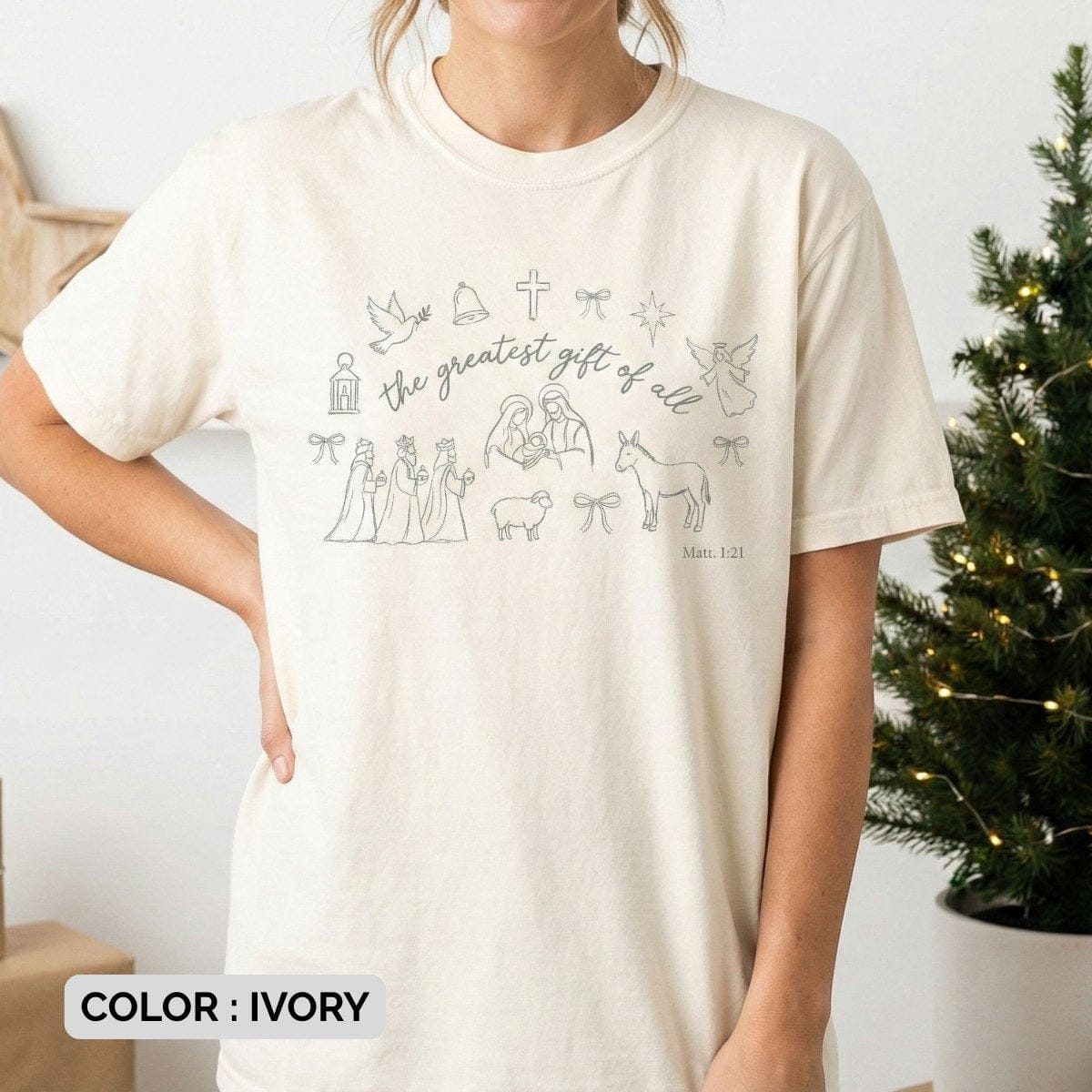 Model wearing an ivory Comfort Colors 1717 Christian women tee featuring minimalist Nativity line art and “the greatest gift of all” Matthew 1:21 design, soft neutral US faith wear styled in a cozy holiday setting.