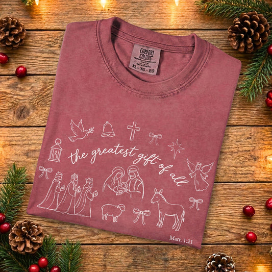 Folded crimson Comfort Colors 1717 Christian women tshirt displaying Nativity scene line art and “the greatest gift of all” scripture design, festive yet minimalist US faith wear on wood background.