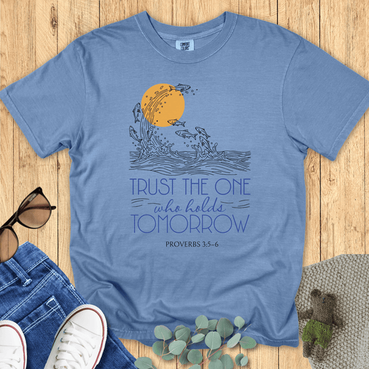 Flat lay of a blue jean Comfort Colors Christian t-shirt with Proverbs 3:5-6 scripture artwork. The print shows fish leaping from waves into the sun, symbolizing faith and trust. Shirt is styled with casual accessories on a wooden background