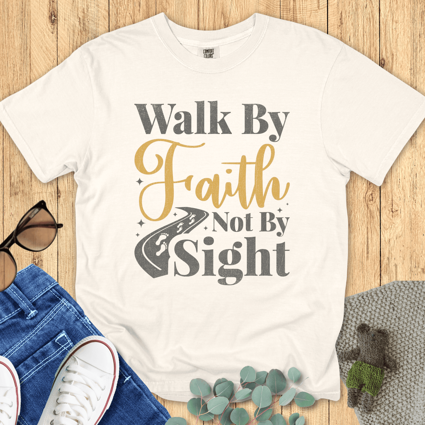 Flat lay of an ivory Comfort Colors Christian t-shirt featuring the scripture-inspired design 'Walk By Faith Not By Sight' with gold and gray typography and a road graphic, styled with jeans, sneakers, sunglasses, and greenery