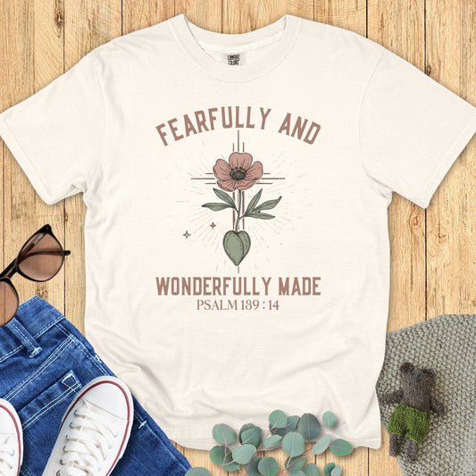 Comfort Colors 1717 ivory “Fearfully and Wonderfully Made – Psalm 139:14” t-shirt flatlay on wooden background with casual outfit pieces. A clean and elegant modern Christian shirt for daily encouragement.