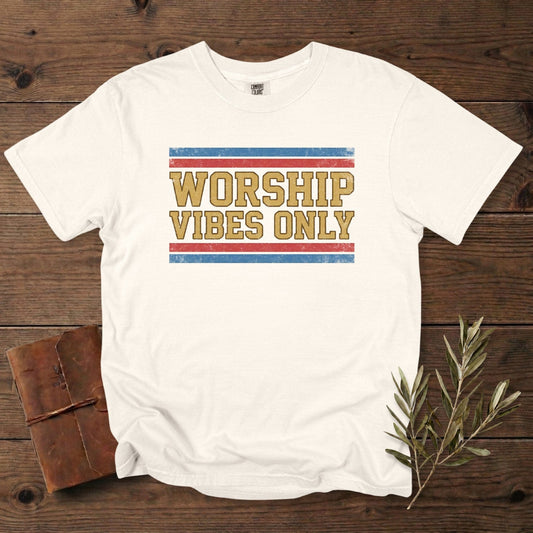Comfort Colors 1717 ivory Christian women tee with a vintage “Worship Vibes Only” text graphic in mustard, red, and blue. Modern Christian tshirt for US faith wear, laid out on a rustic wooden background with journal and olive leaves.