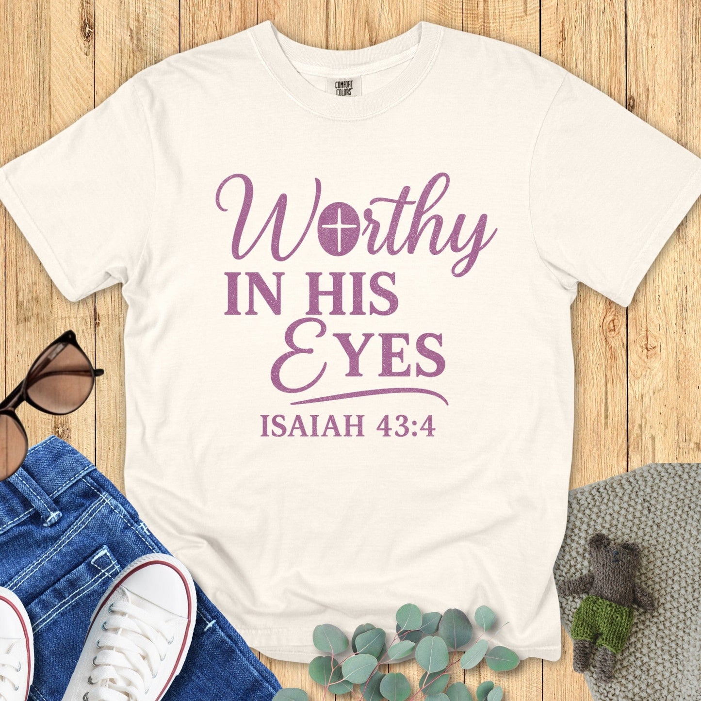 Comfort Colors 1717 ivory Christian t-shirt with the phrase 'Worthy in His Eyes – Isaiah 43:4' in purple lettering, displayed in a styled flat lay with denim jeans, sneakers, and greenery accents.